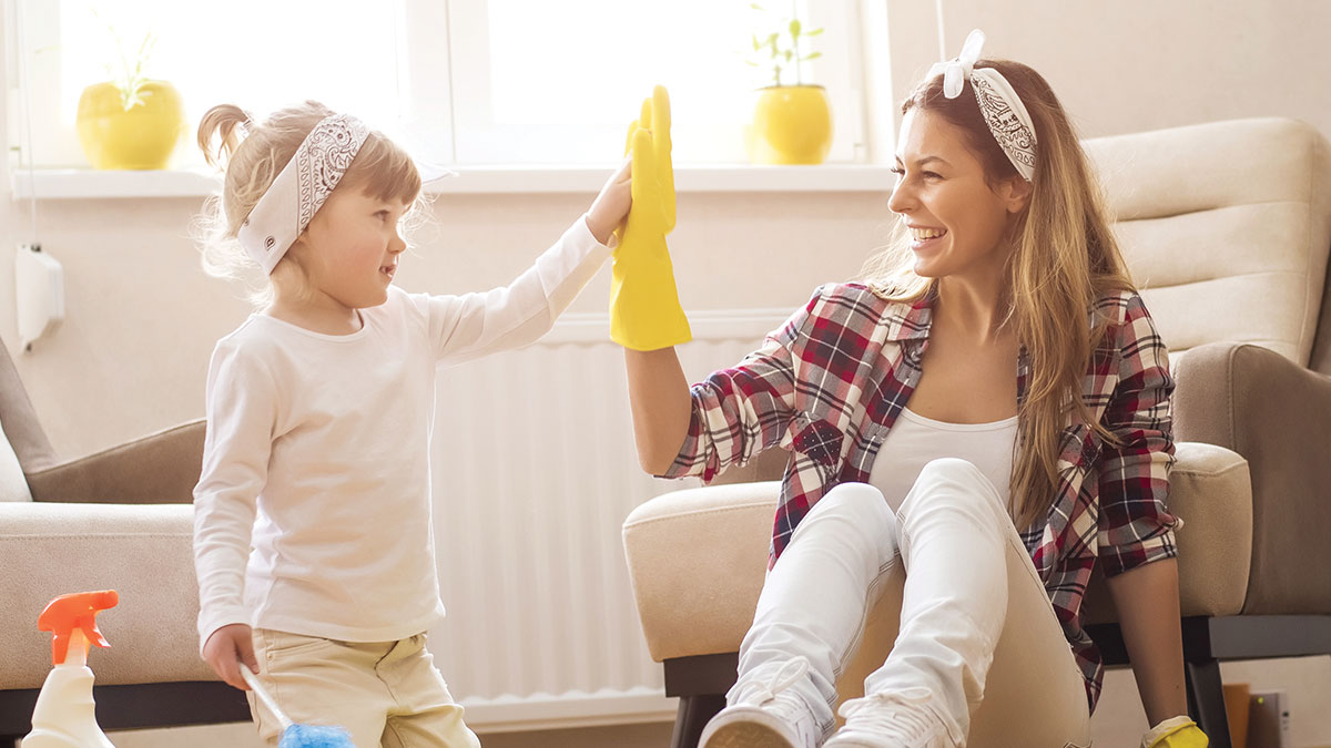 Parent and child share a high five while tidying a bright living room, illustrating a friendly, family-focused approach to home care.