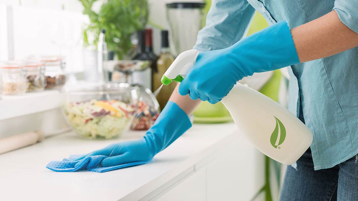 Gloved hands spray and wipe a kitchen countertop during routine cleaning, showing careful surface-by-surface work.